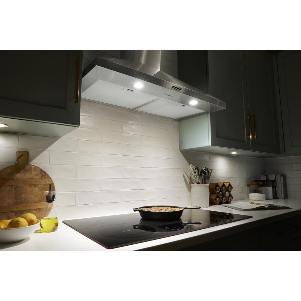 Stainless steel chimney range hood with LED lights above a modern black glass cooktop in kitchen and appliances setting