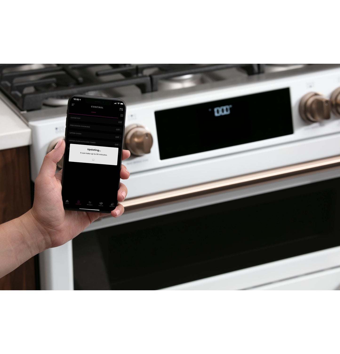Hand holding smartphone updating app controlling white smart gas double-oven range with bronze knobs from Ranges collection