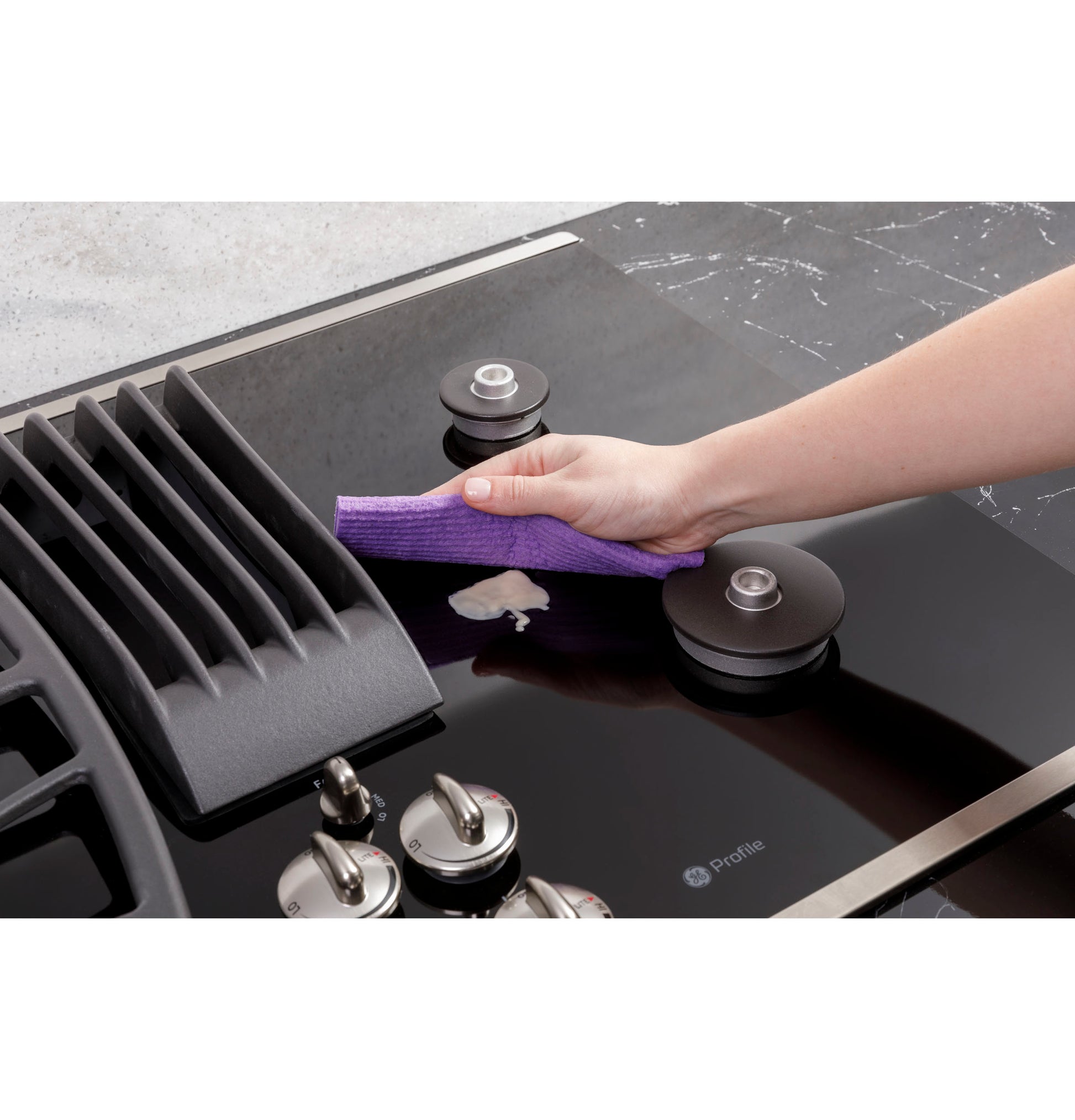 Person wiping glossy black gas cooktop with downdraft vent and stainless steel knobs in modern kitchen