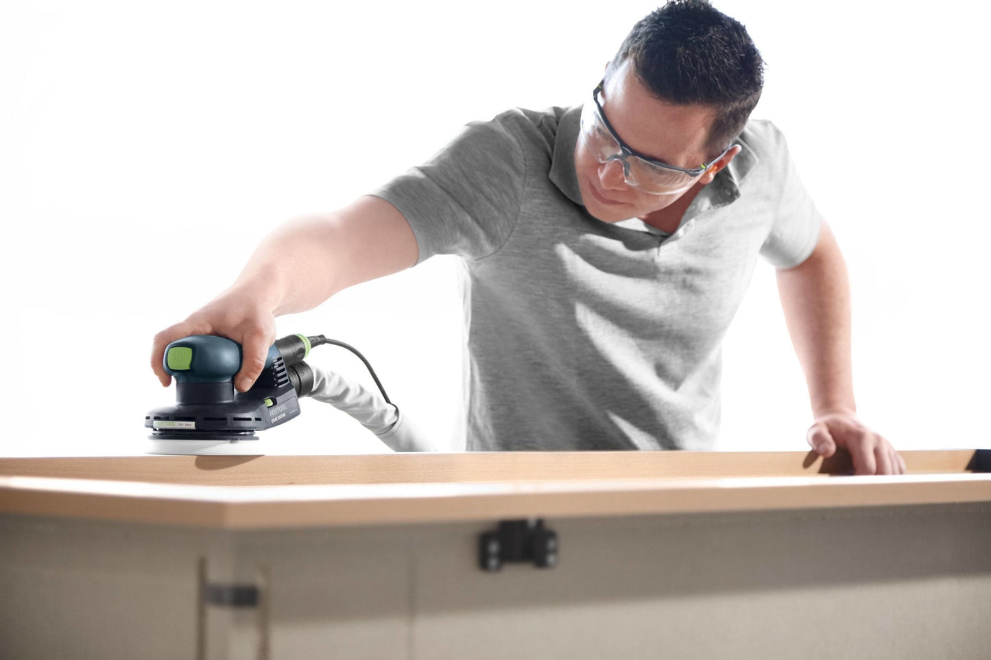 Man using a random orbit sander to smooth wooden furniture in a bright workshop setting