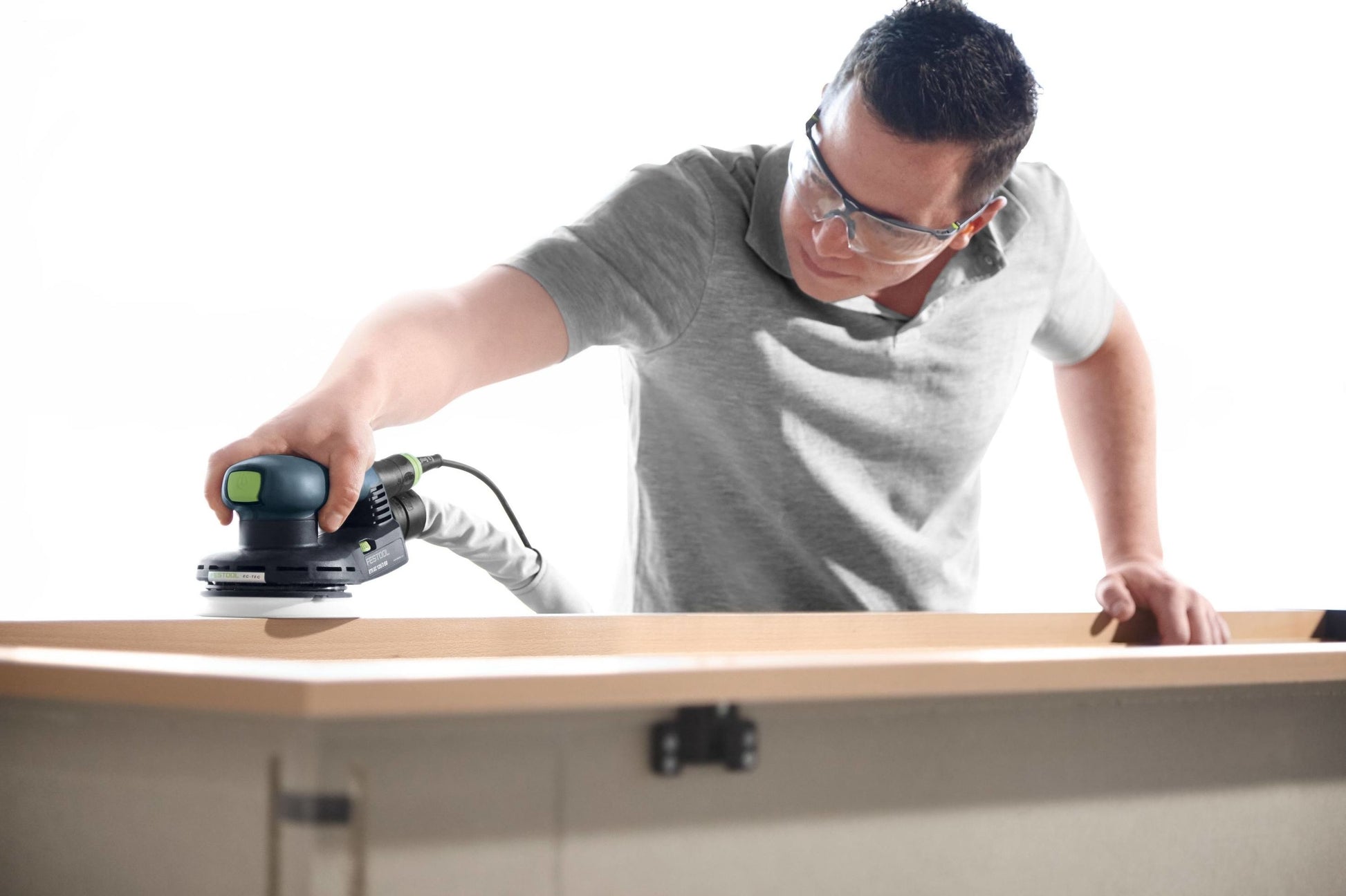 Man using a random orbit sander to smooth wooden furniture in a bright workshop setting