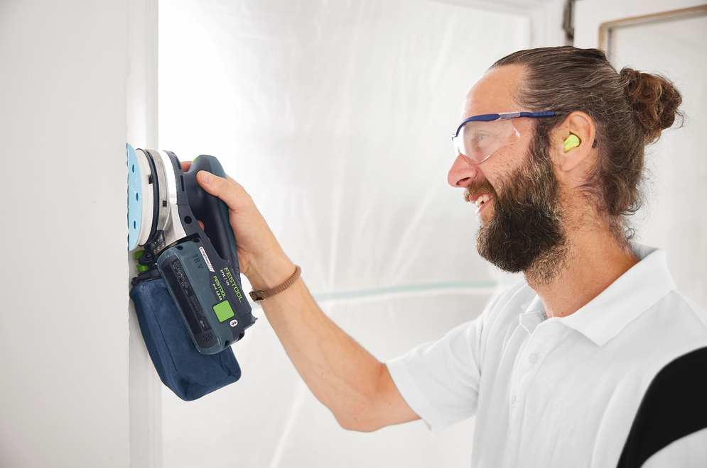 Man wearing safety glasses and earplugs using a Festool cordless random orbit sander on white wall in workshop