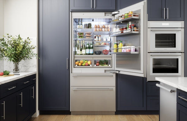 Built-in stainless steel refrigerator with bottom freezer integrated into navy kitchen cabinetry and stocked interior shelves