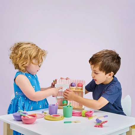 Two children playing with a wooden ice cream kiosk and colorful pretend food on a white table