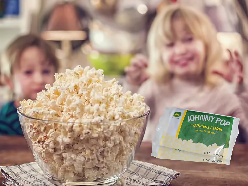 Clear glass bowl filled with freshly popped popcorn on a wooden table with two children eagerly reaching for it in the background.