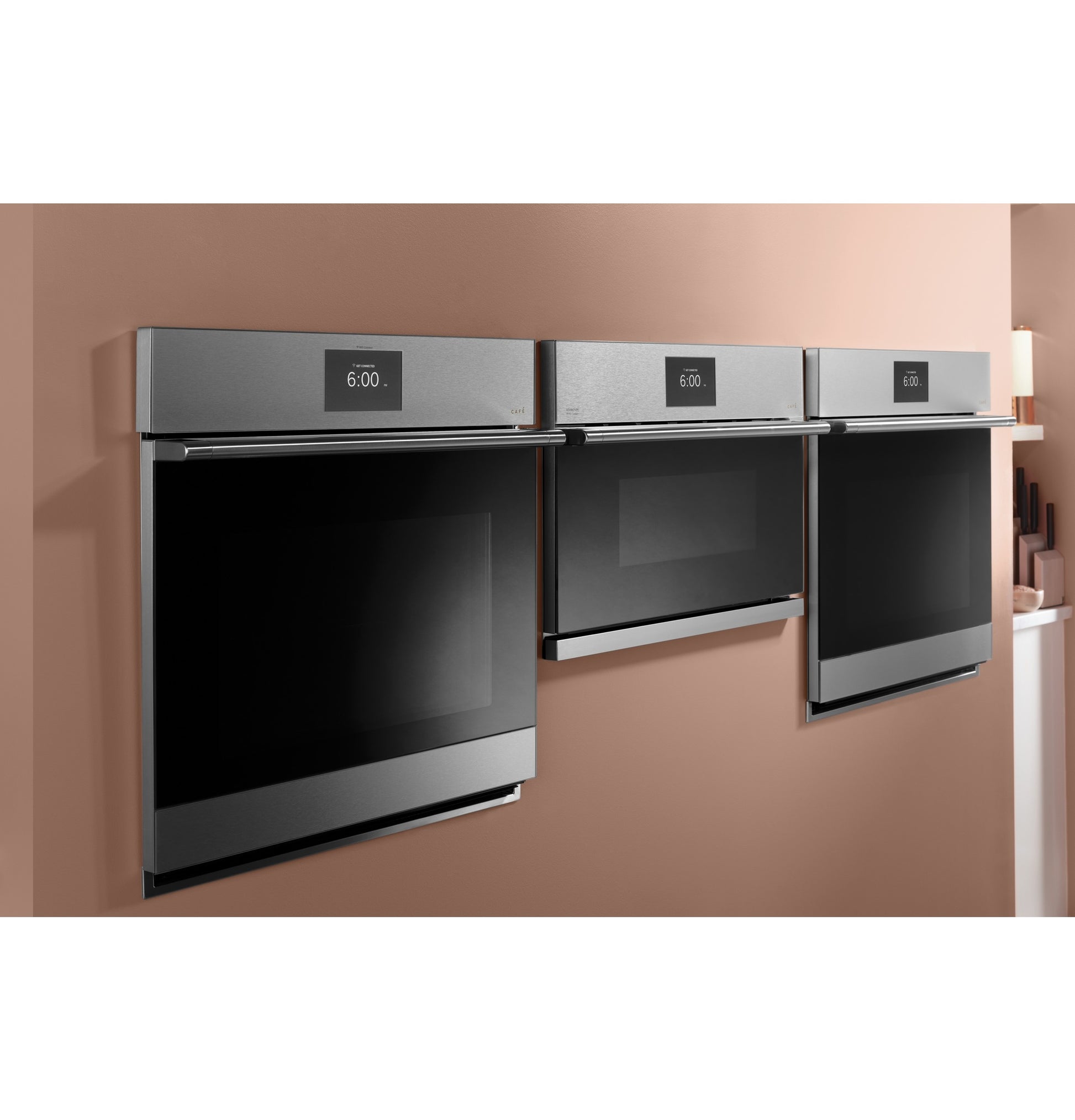 Three sleek stainless steel wall ovens with digital clocks mounted on a warm-toned kitchen wall in appliance setting