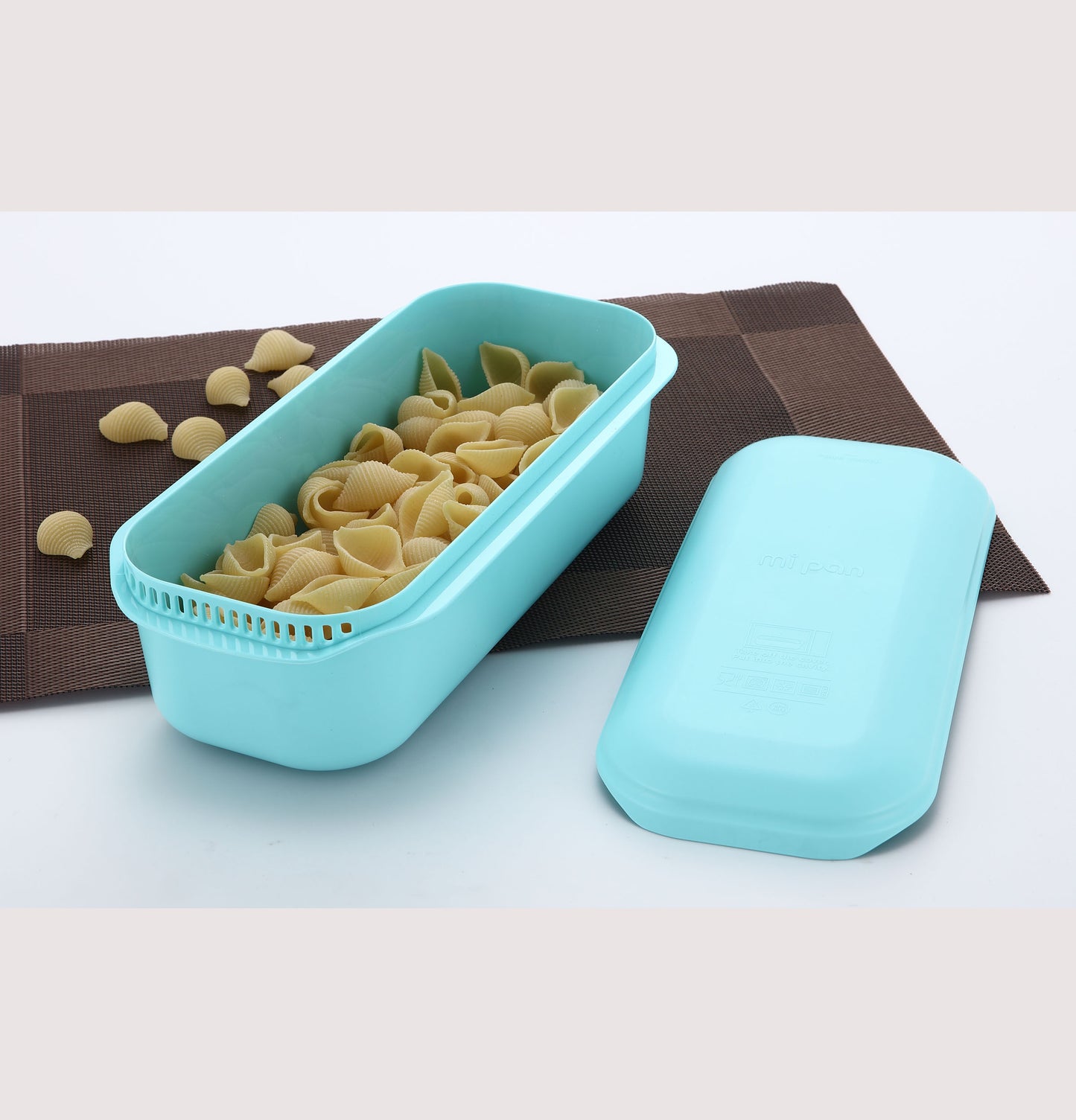 Blue microwave pasta cooker with uncooked shell pasta on brown placemat in kitchenware collection
