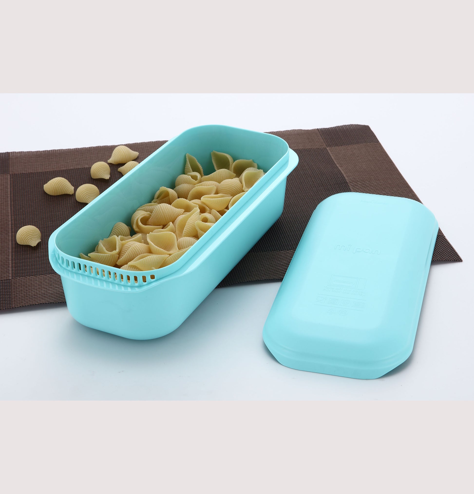 Blue microwave pasta cooker with uncooked shell pasta on brown placemat in kitchenware collection