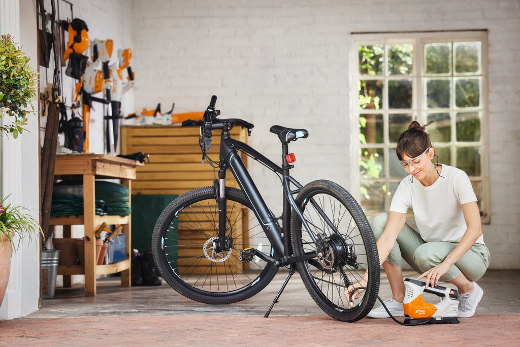 Person using a compact battery-powered inflator to air up a black bicycle tire in a bright home workshop, part of Power Tools and Air Compressors collections.