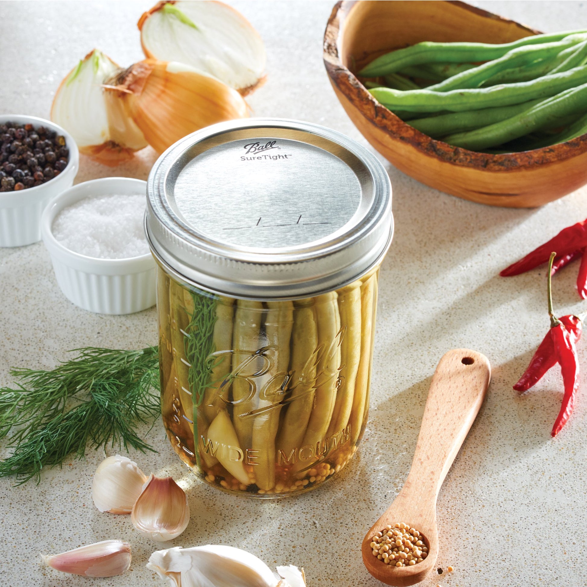 Clear ball wide mouth canning jar filled with pickled green beans surrounded by garlic, onion, spices, and herbs