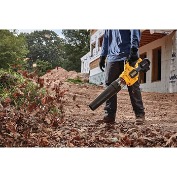 Person wearing gloves using a yellow cordless axial blower outdoors to clear leaves at a construction site
