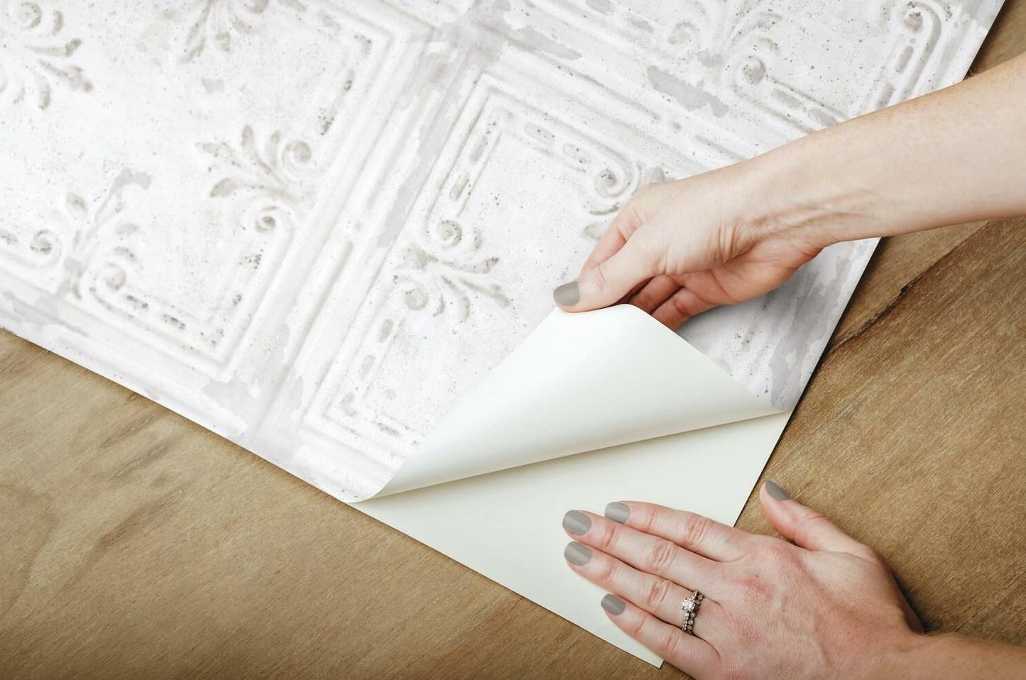 Hands peeling backing from white embossed tin tile patterned peel-and-stick wallpaper on wooden surface