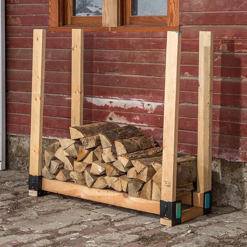 Wooden log stacker with metal brackets holding split firewood against rustic red wooden wall outdoors