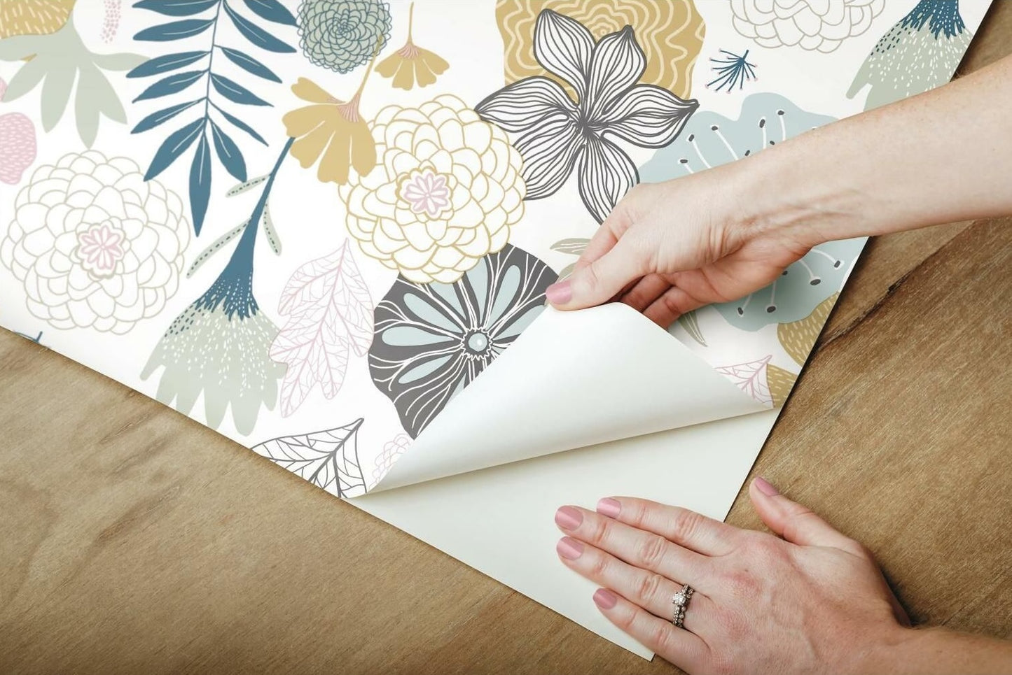 Hands applying pink and beige floral peel and stick wallpaper with leafy accents on wooden surface