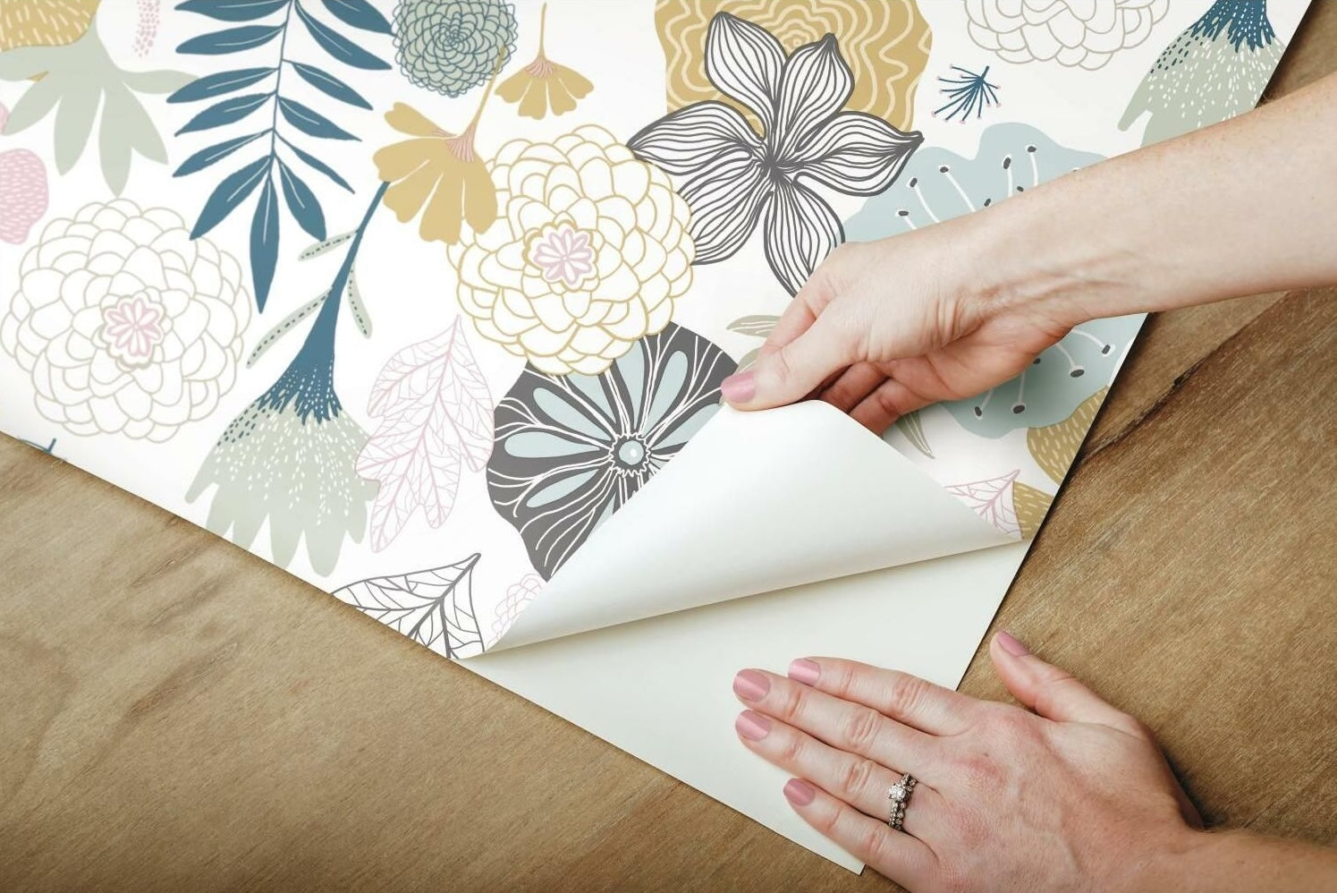 Hands applying pink and beige floral peel and stick wallpaper with leafy accents on wooden surface