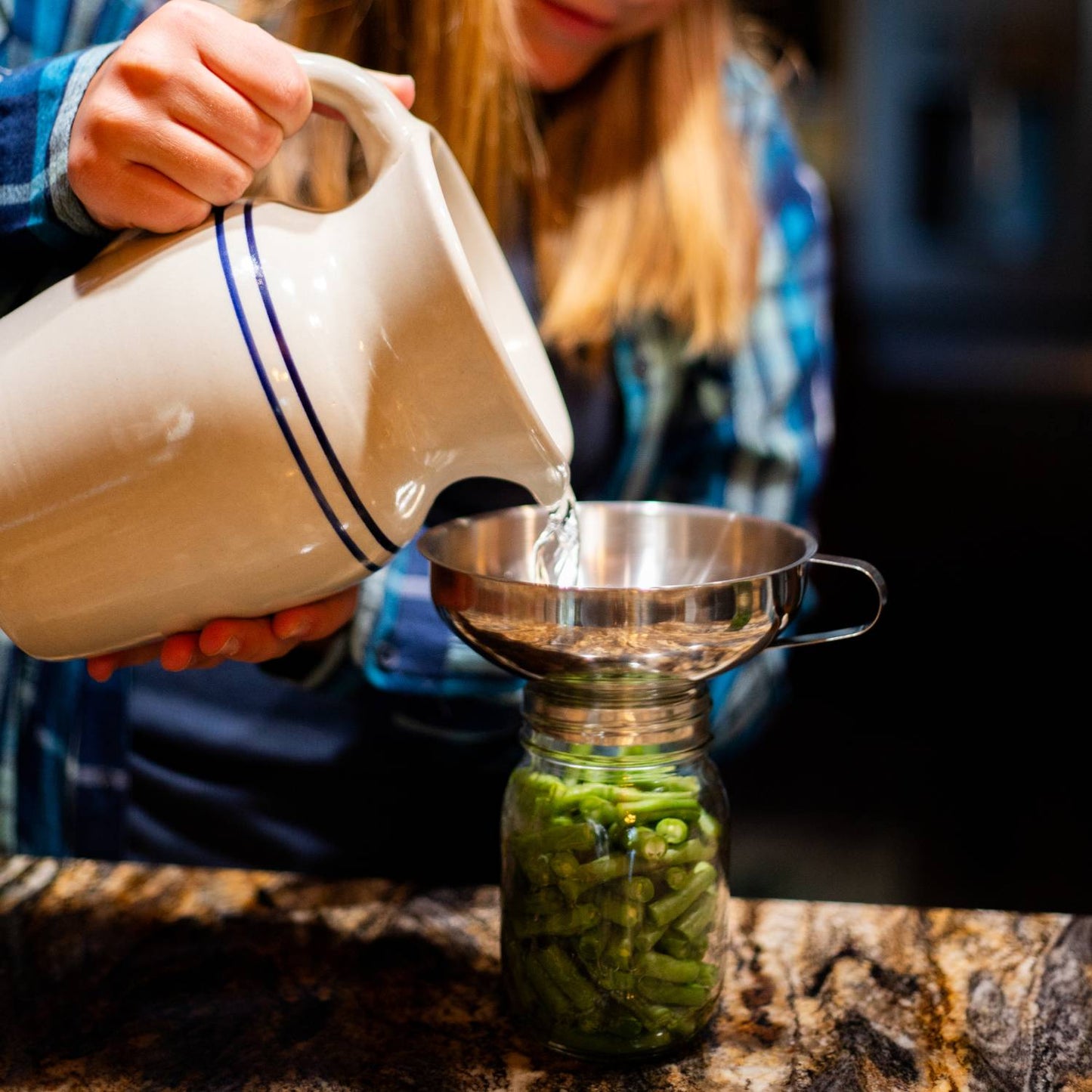 Person pouring liquid from ceramic pitcher into stainless steel funnel filling jar with green beans in kitchenware collection
