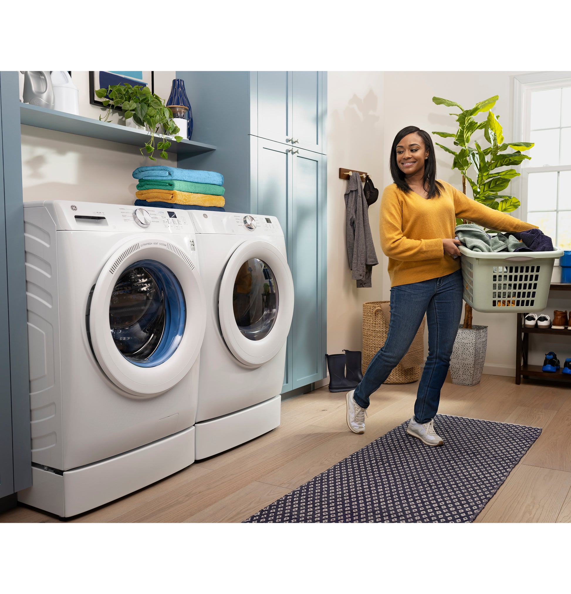 Woman carrying laundry basket near white front load washer and dryer in modern laundry room with blue cabinets