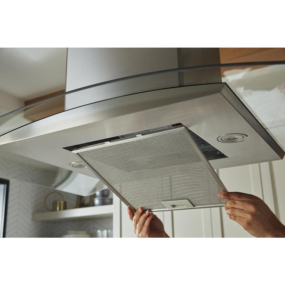 Hands removing metal grease filter from stainless steel curved glass kitchen island vent hood in modern kitchen