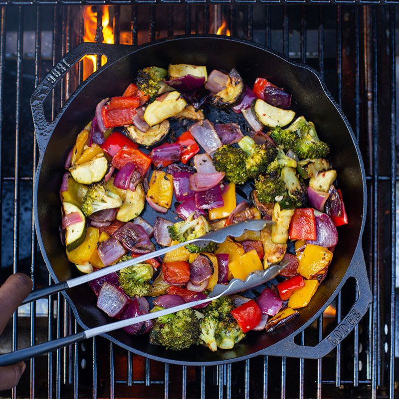Seasoned cast iron skillet with dual handles grilling colorful vegetables over open flame in kitchenware and Lodge Cast Iron collections