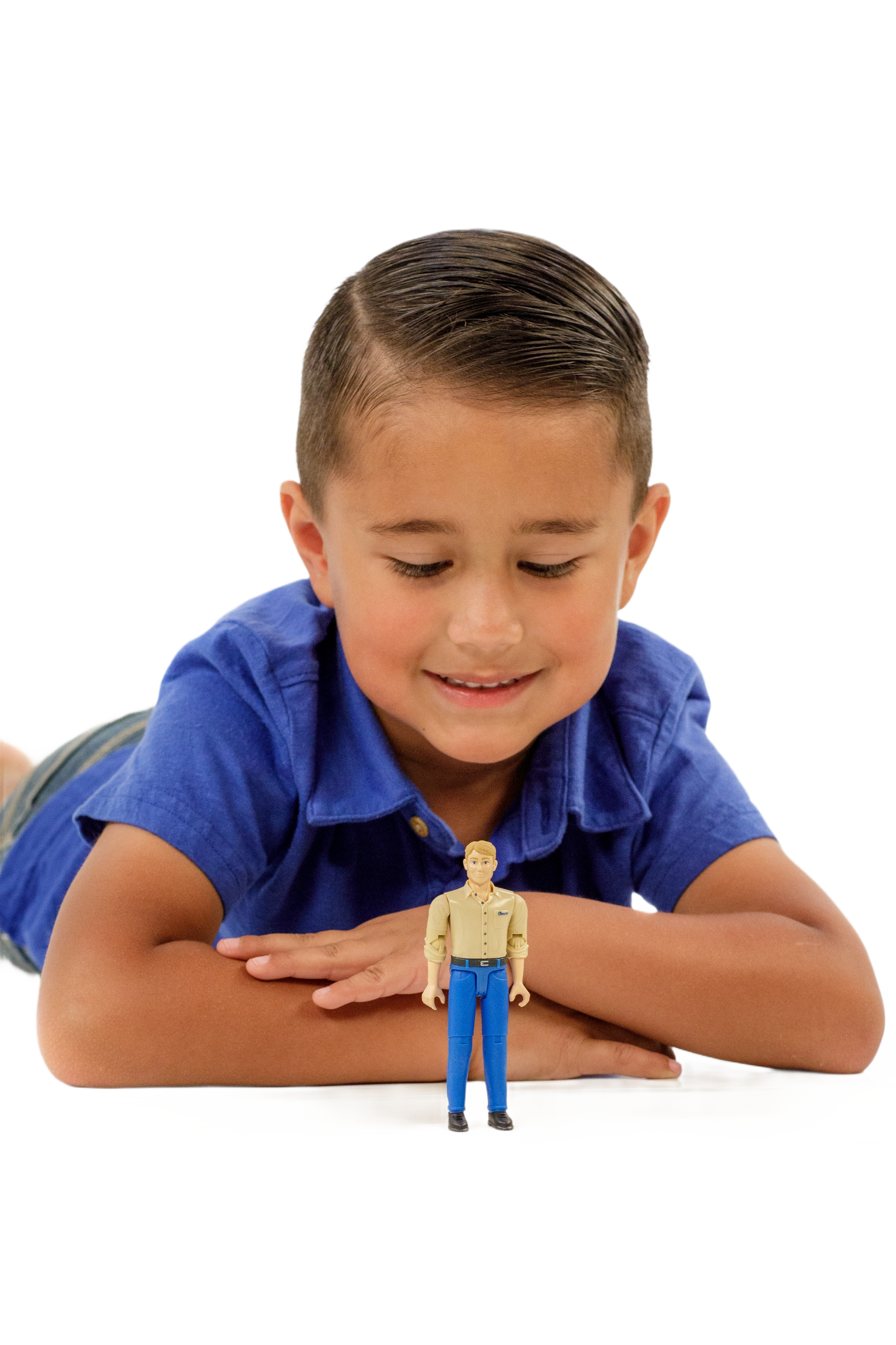 Child lying down smiling at a light skin action figure dressed in blue pants and tan shirt on white surface