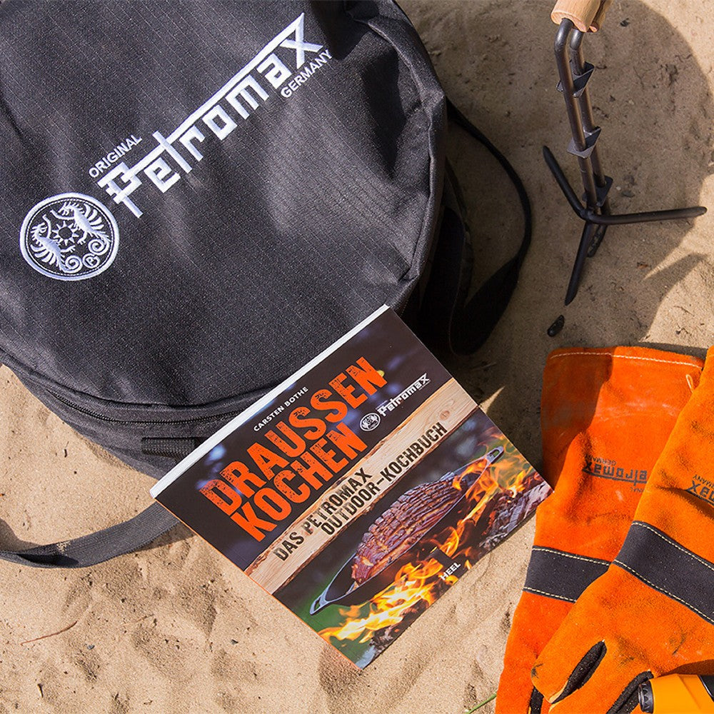 Black Petromax transport bag with outdoor cooking book and orange heat-resistant gloves on sandy surface