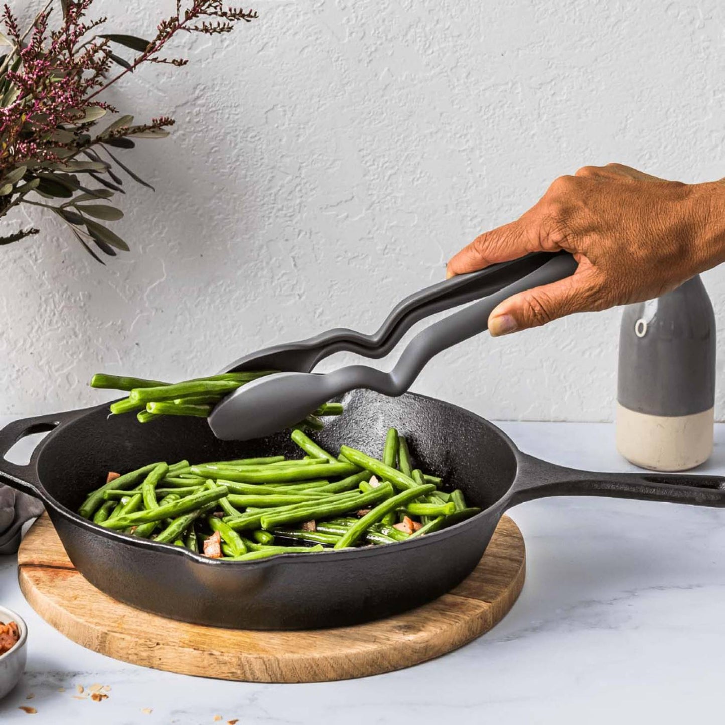 Hand holding gray 10.5-inch kitchen tongs picking green beans from a cast iron skillet on wooden board in kitchenware setting