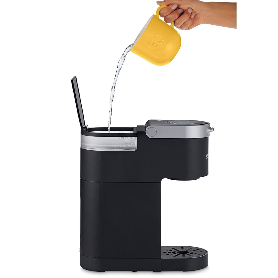 Black single-serve coffee machine with silver accents being filled with water from a yellow measuring cup for home use