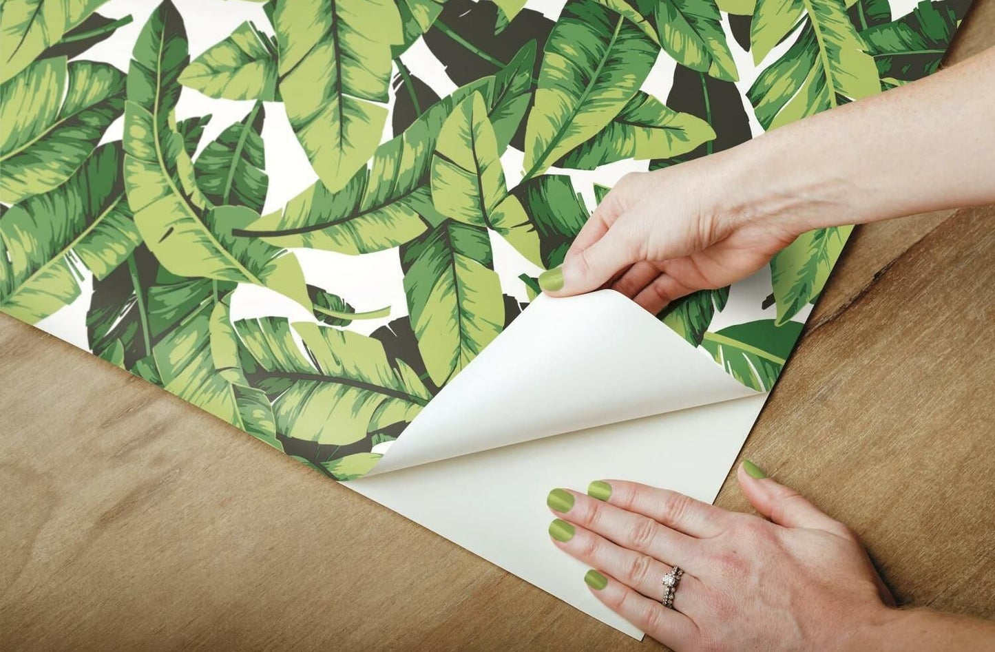 Hands peeling green palm leaf patterned peel and stick wallpaper from backing paper on wooden surface