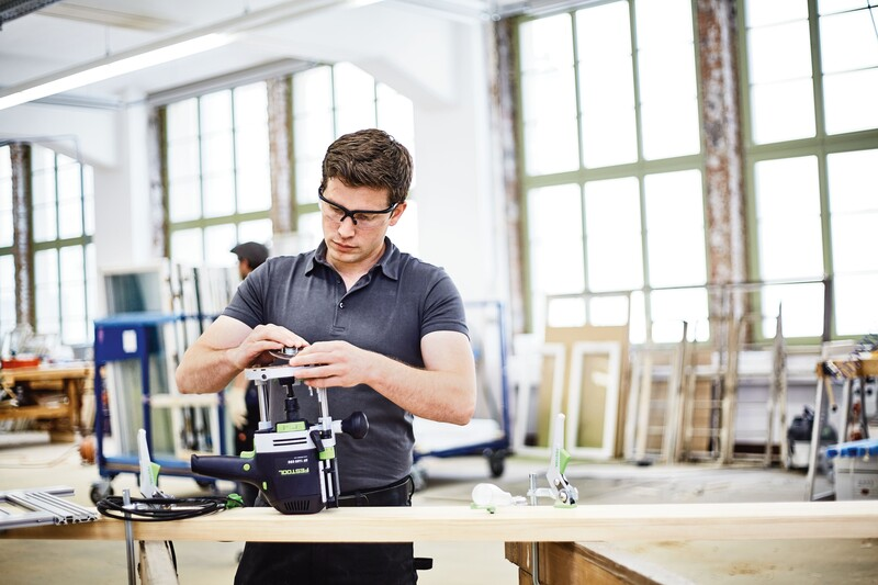 Man adjusting a Festool router tool in a bright woodworking shop with large windows, part of woodworking accessories.