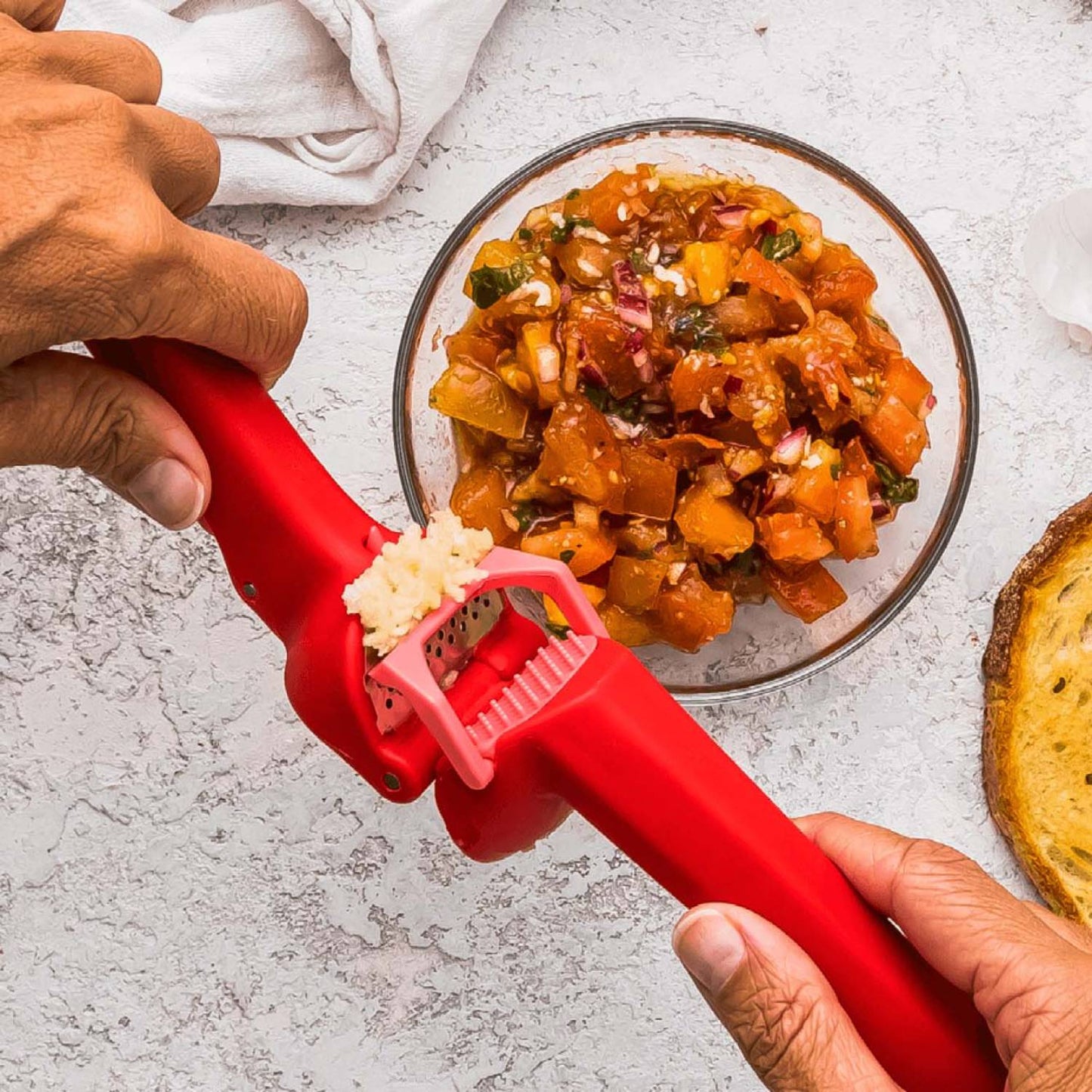 Red garlic press with ergonomic grip crushing fresh garlic over a bowl of homemade salsa in kitchen utensils collection