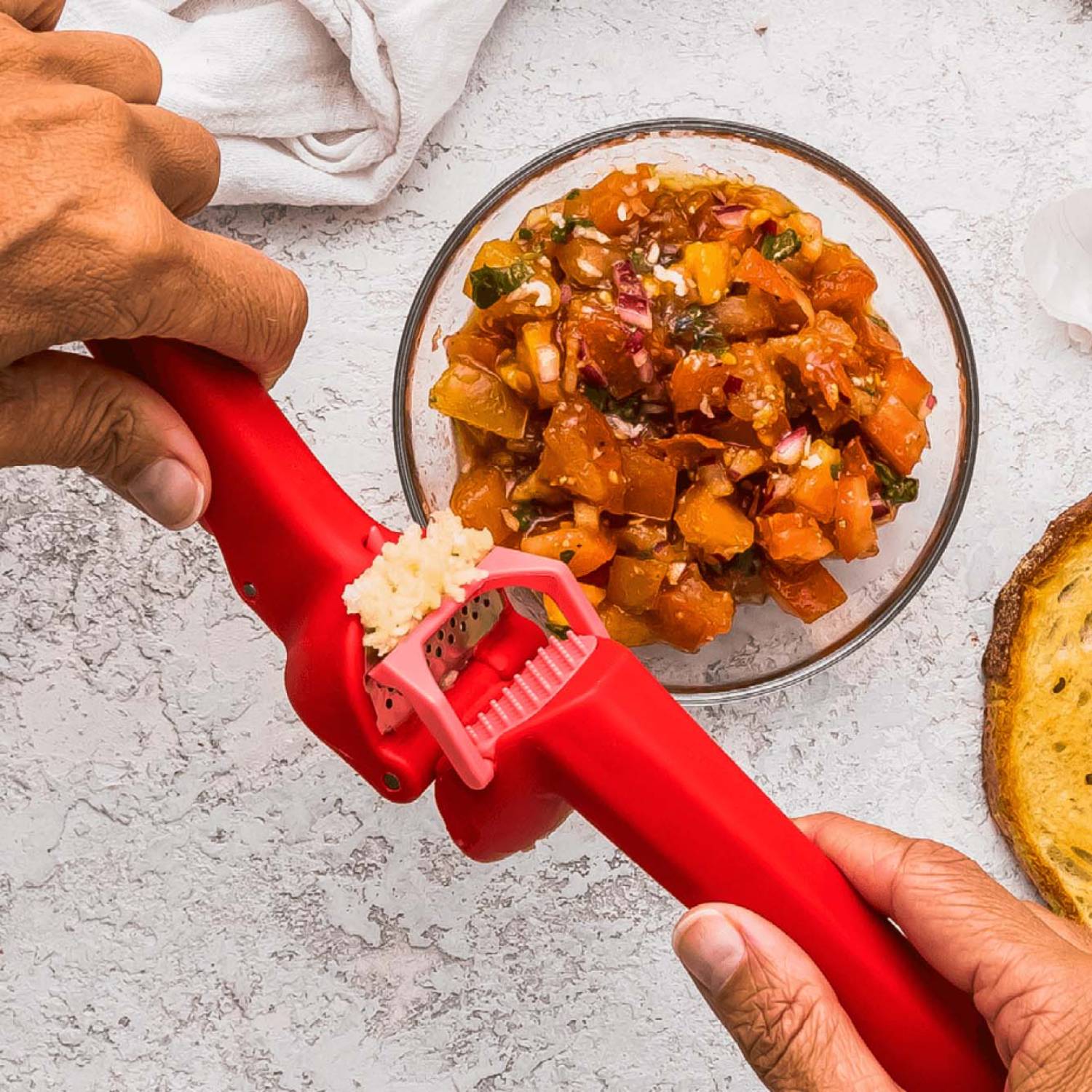 Red garlic press with ergonomic grip crushing fresh garlic over a bowl of homemade salsa in kitchen utensils collection