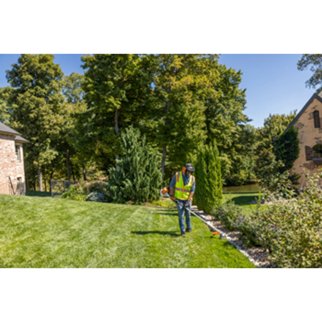 Person wearing safety vest trimming edge of lush green lawn near decorative shrubbery and stone walkway for home improvement task