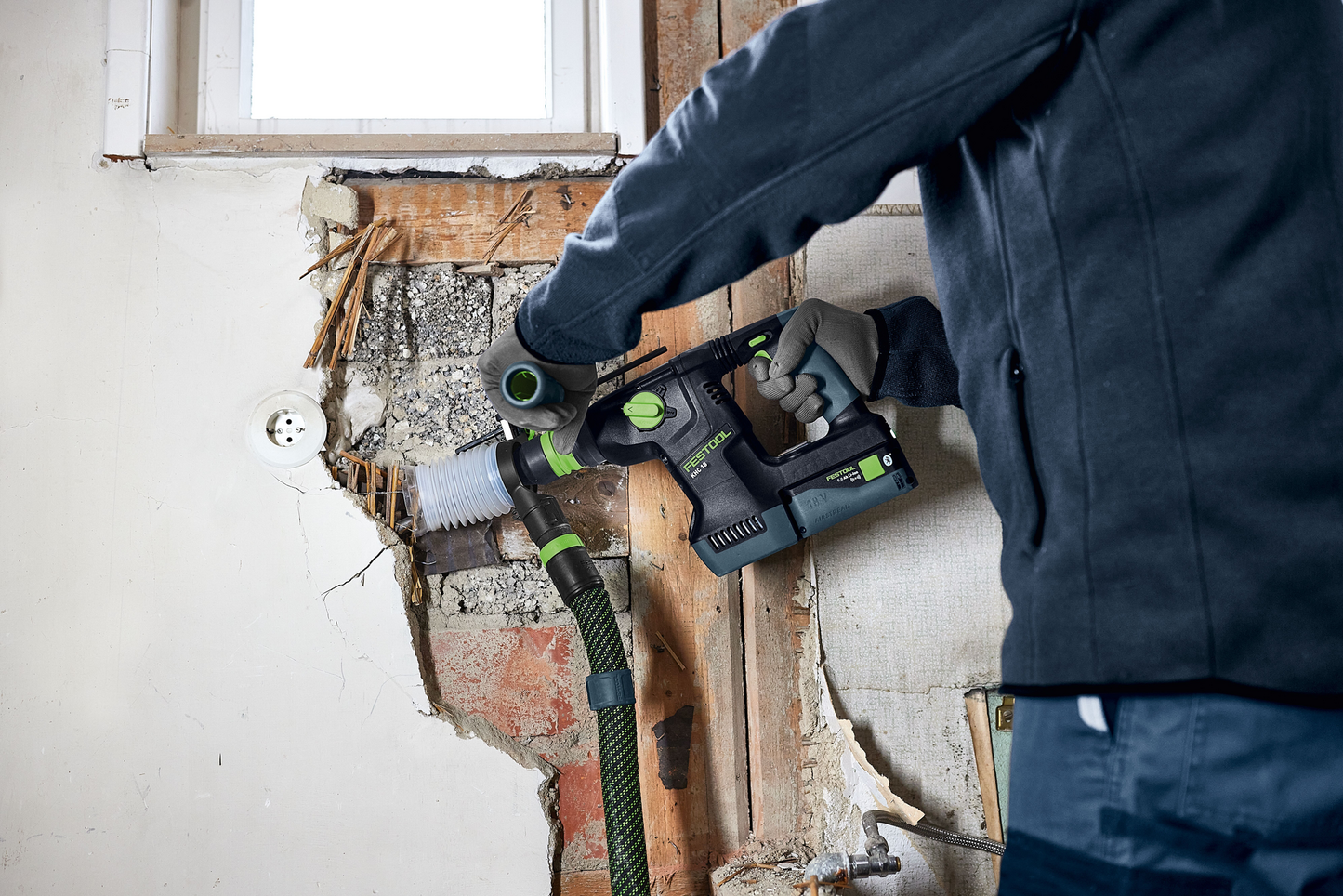 Black and green drill with auxiliary handle attached, used for precise chiseling on damaged indoor wall surface in home improvement