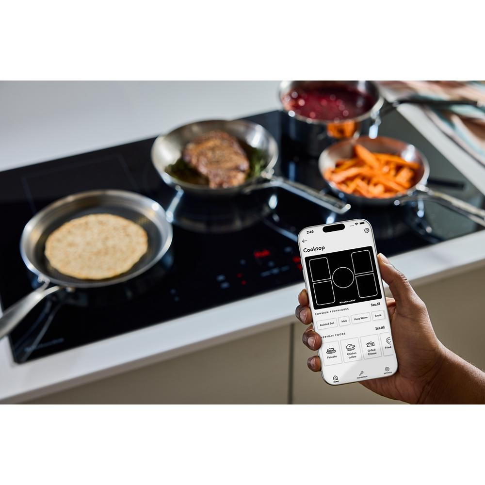 Person holding smartphone displaying cooktop controls near black induction cooktop with pans cooking food in modern kitchen