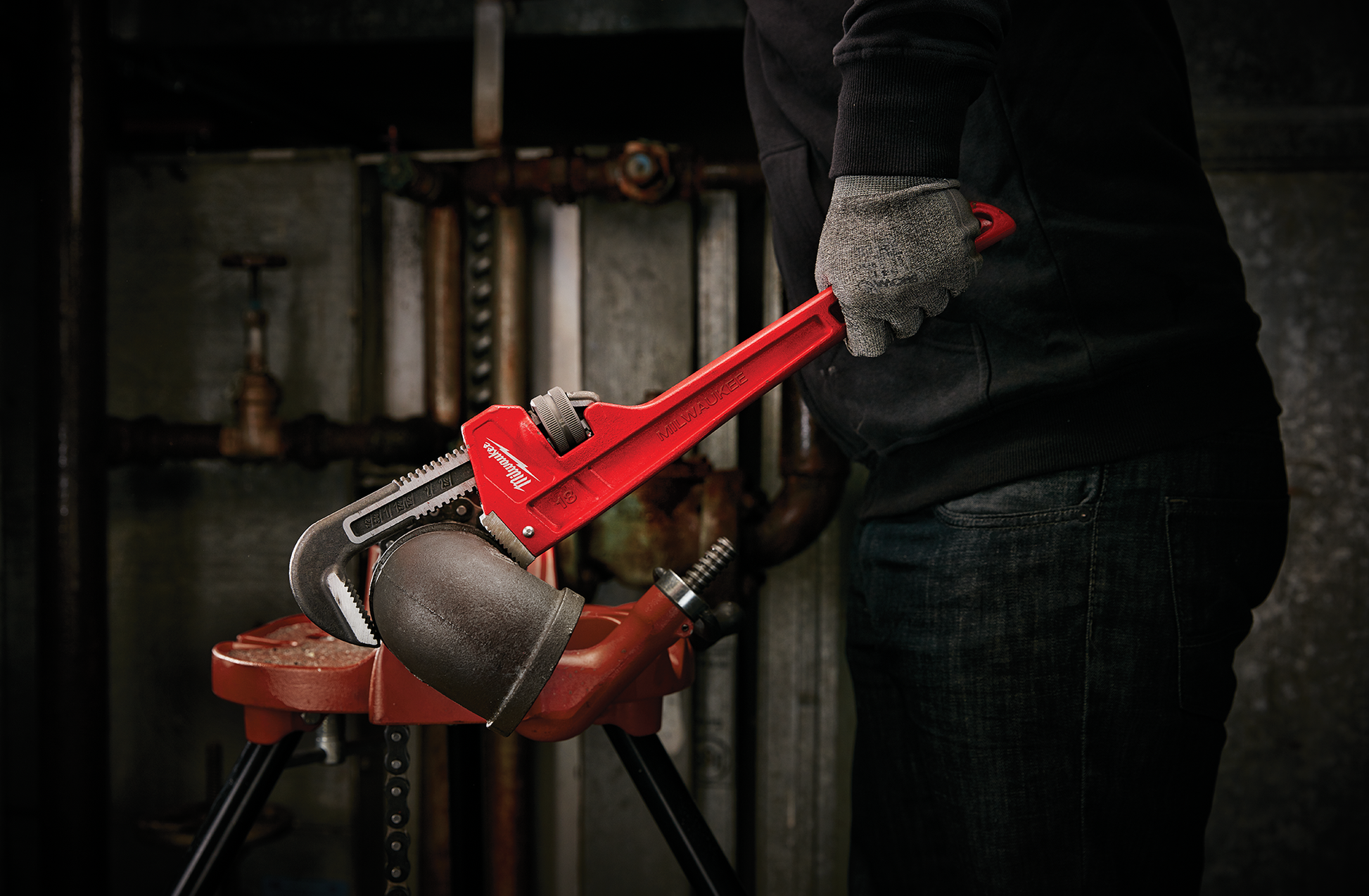Red steel pipe wrench gripping a black elbow pipe on a red pipe vise in a plumbing workshop setting