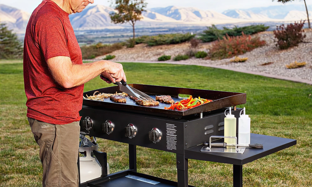Man cooking seasoned meat and sliced bell peppers on a large outdoor flat-top grill set on a grassy yard with mountains in the distance.