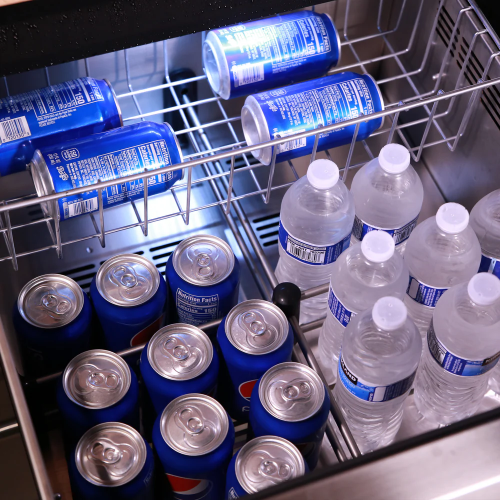 Open beverage cooler drawer with blue soda cans and plastic water bottles under bright interior LED lighting