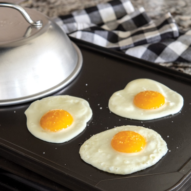 Aluminum dome with handle resting on griddle with three cooked sunny-side-up eggs for kitchen gadgets and tools