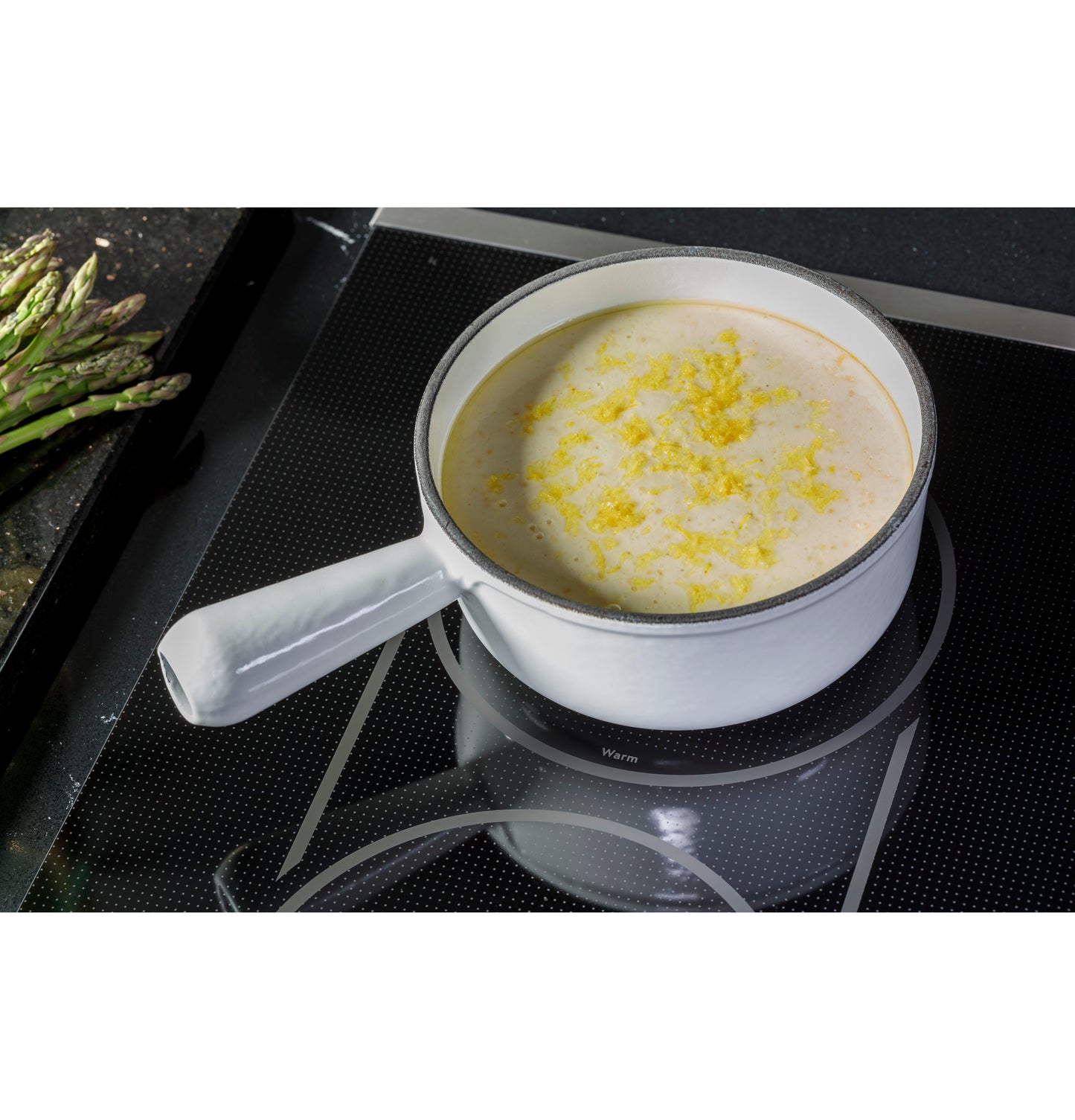 White saucepan cooking creamy sauce on a black glass touch-control cooktop with asparagus nearby in kitchen setting