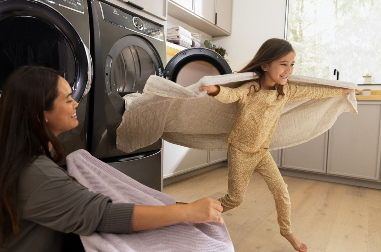 Mother folding laundry beside black front load electric dryer in bright kitchen with child playing nearby in pajamas