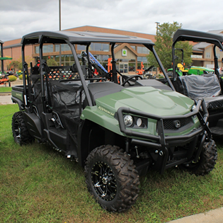 Green utility vehicle with four seats and rugged off-road tires parked on grass near buildings