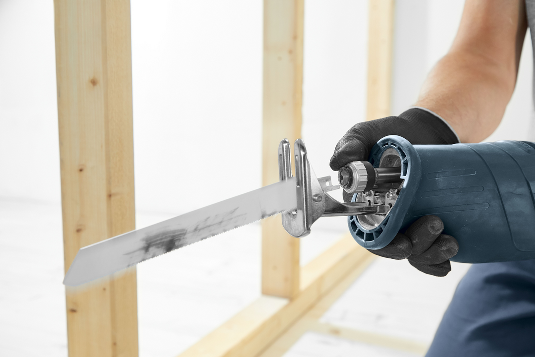 Person gripping blue cordless reciprocating saw with black gloves near wooden frame under construction