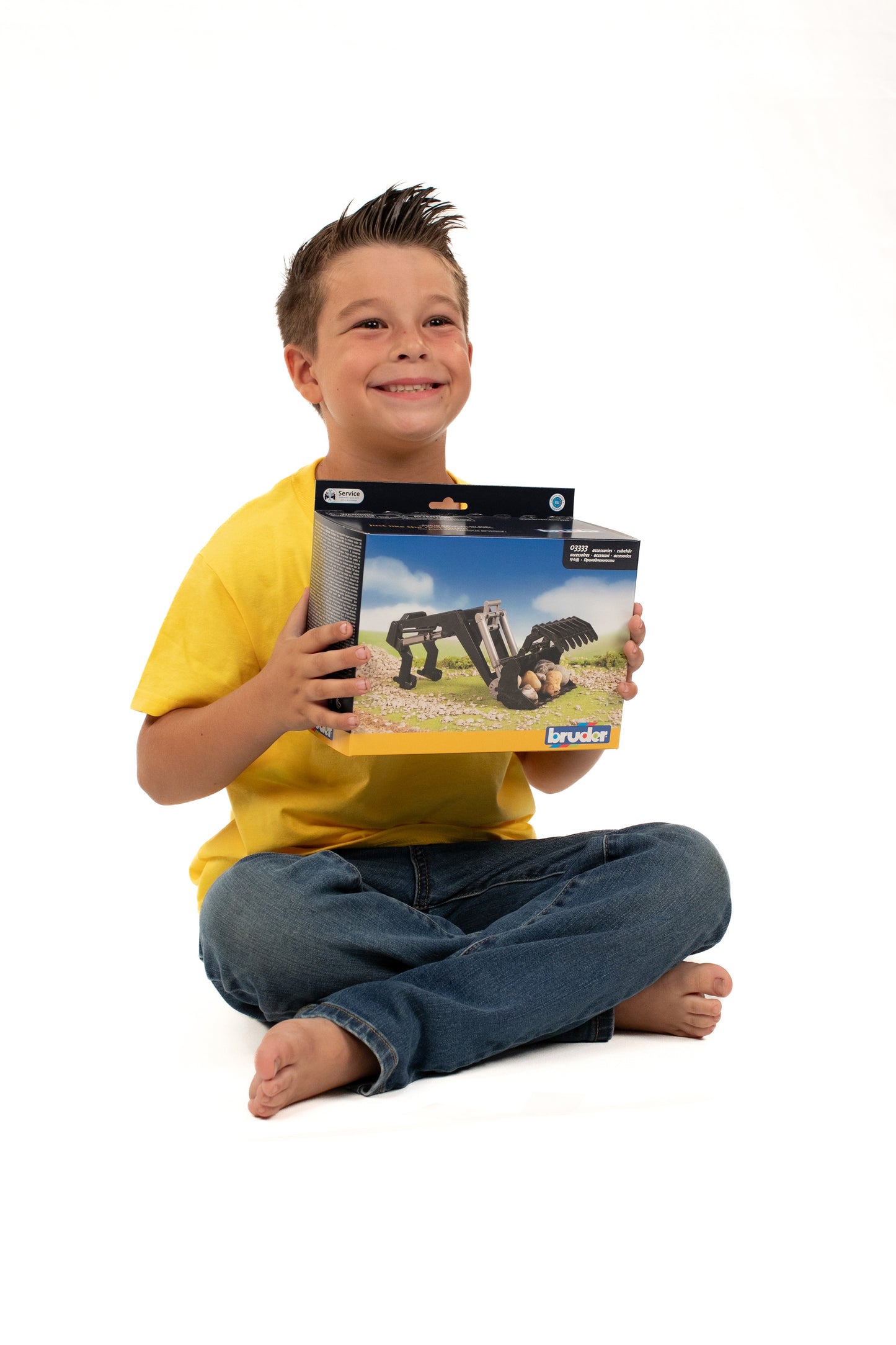 Smiling boy in yellow shirt sitting cross-legged holding Bruder front loader toy packaging for home play and tools improvement