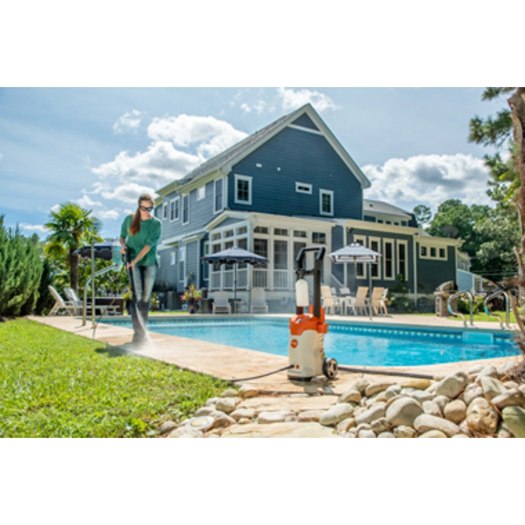 Person using electric pressure washer with hose and spray wand to clean grass near backyard pool and large home