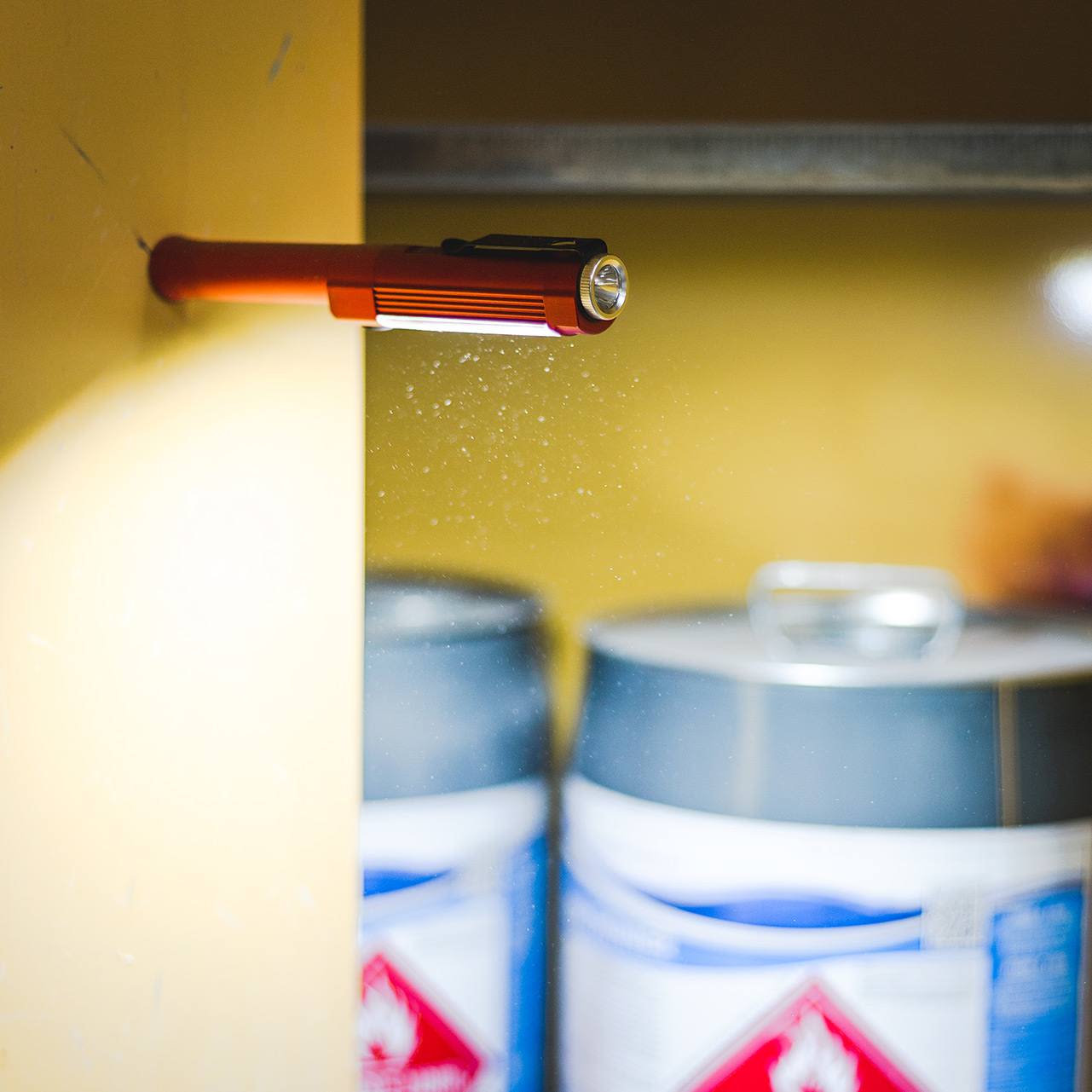 Red power work flashlight with magnetic base illuminating a yellow metal wall near industrial chemical barrels