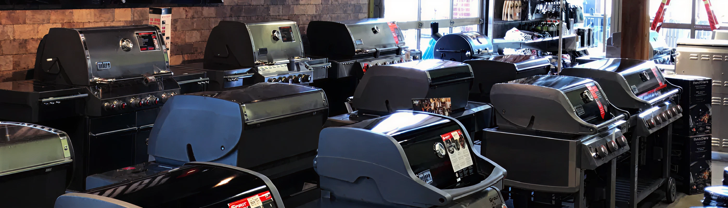 Indoor showroom with multiple closed barbecue grills arranged in rows near a large window, featuring brick walls and natural light.