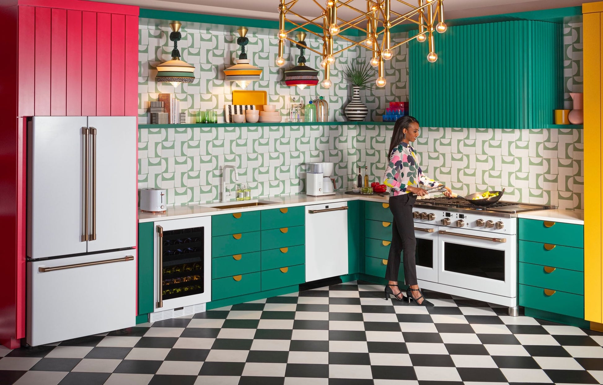 Bright retro kitchen with teal and red cabinets, checkered floor, gold chandelier, and woman cooking at a stove