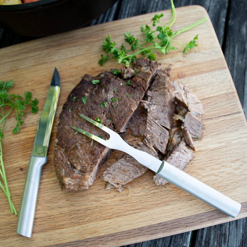 Two-pronged carving fork with brushed handle beside sliced steak and fresh herbs on wooden cutting board in kitchenware collections