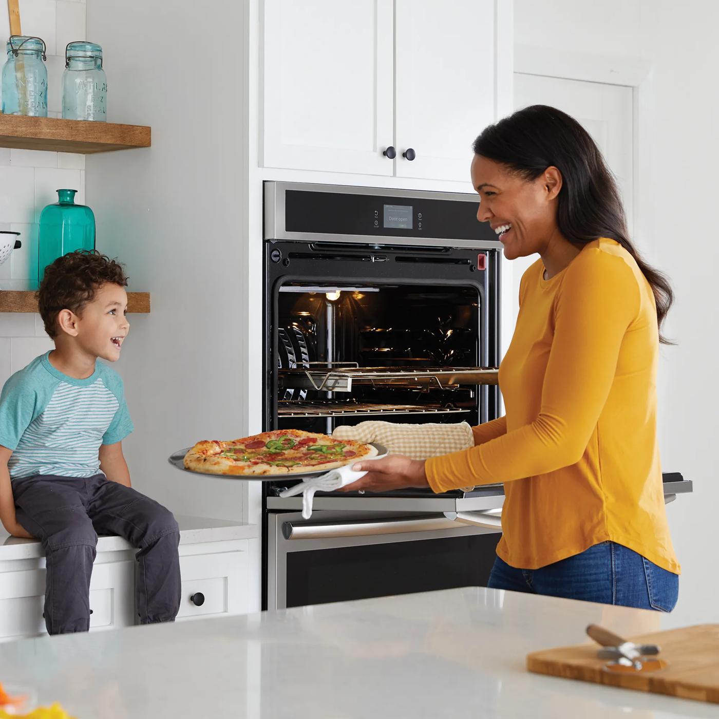 Woman removing pizza from built-in double electric wall oven while child watches in modern kitchen setting