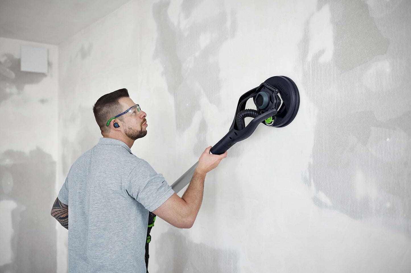 Man wearing safety glasses using a drywall sander on a patched wall in a home improvement setting